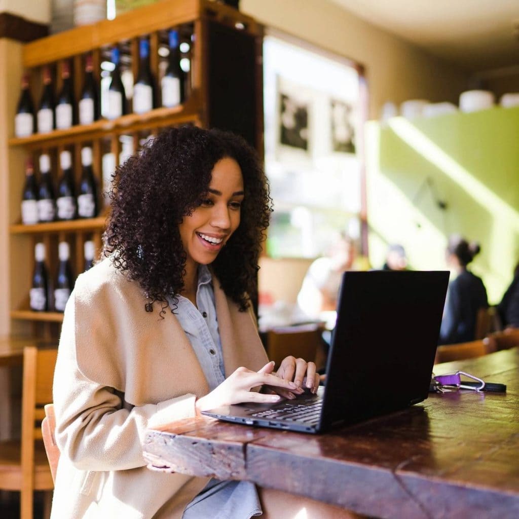 Young woman on her laptop in her wine shop, excitedly checking the current schedule of AI classes on savageai.com, making an informed decision as an entrepreneur.